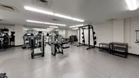 This fitness center is a fully outfitted exercise facility designed to support a wide range of workout routines. The space is laid out across a single floor with a practical, open floor plan that maximizes usability and flow between equipment zones. The interior features clean white walls, light-toned flooring with durable fitness-grade tiles, and bright overhead fluorescent lighting that ensures the space is well-lit throughout.

The cardio section is prominently featured, with a row of modern treadmills equipped with individual display screens, alongside an upright stationary bike. Full-length mirrors line one wall, visually expanding the space and allowing users to monitor their form during exercise. A wall-mounted television provides additional entertainment for cardio sessions.

The strength training area is equally well-appointed, featuring a power rack with weight plates, a cable machine, a leg press or seated machine, an adjustable bench, and a dumbbell rack with a range of free weights. This variety of equipment supports both machine-based and free weight training routines.

A hydration station is conveniently located near the entrance, featuring a dual-level stainless steel drinking fountain alongside a wall-mounted hand sanitizer dispenser. The overall ambiance of the fitness center is clean, functional, and professional, making it a comprehensive facility suited for a broad range of fitness needs.