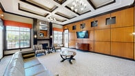 Located at 900 Grand Central Dr in Hamilton, NJ, this sophisticated single-floor clubhouse and leasing lounge offers a generously proportioned, open-plan gathering space designed with upscale finishes throughout. The space is anchored by striking coffered ceilings adorned with elegant chandeliers, lending a sense of architectural grandeur. Warm honey-toned wood wall paneling dominates one side of the main lounge area, featuring recessed display niches and a wall-mounted flat-screen television above a classic demilune console. A sleek reception desk greets visitors upon entry, with comfortable blue lounge seating nearby. A cozy fireplace surrounded by mosaic tile work and a decorative mirror serves as a focal point in the sitting area, which is furnished with patterned accent chairs and a glass-topped coffee table. Large windows dressed with bold orange valances flood the interior with natural light and offer views of the outdoor pool area. Transitioning into the café zone, hardwood flooring, vibrant pendant lighting, mosaic tile backsplash, and bar-height counter seating create a casual yet stylish atmosphere. The cohesive design palette of dark wood trim, warm neutrals, and rich textures gives the entire space a refined, welcoming ambiance ideal for residents and guests alike.