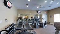 This fitness center is designed to provide a comprehensive workout environment within a single, open-layout floor. The space is outfitted with a lineup of cardio equipment, including multiple treadmills and elliptical machines, arranged in rows for easy access and ample movement between stations. A dedicated free weight area features a rack of dumbbells, and a Hoist weight bench adds strength-training capability to the room. A large wall-length mirror runs along one side of the space, enhancing the sense of openness and allowing users to monitor their form during exercise. The walls are finished in a neutral warm tone, contributing to an energizing yet comfortable atmosphere. Recessed lighting illuminates the room evenly, complemented by a ceiling fan that helps maintain airflow throughout workouts. A wall-mounted flat-screen television is positioned for visibility across the floor, providing entertainment during cardio sessions. An exit sign and emergency access door are clearly marked, reflecting attention to safety standards. The flooring includes both rubber mat sections under equipment and carpeted areas, offering both durability and comfort. Natural light filters in through windows visible in the mirror's reflection, adding brightness to the overall environment.
