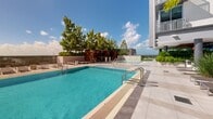 Located at 9600 S Dixie Hwy in Miami, FL, this amenity level showcases a thoughtfully designed outdoor retreat that blends South Florida's tropical character with modern architectural sensibility. The centerpiece is a large resort-style swimming pool flanked by generous sun decks finished in large-format porcelain tile, lined with contemporary lounge chairs and seating clusters ideal for relaxation. Adjacent to the main pool sits a dedicated spa featuring dramatic water-jet fountains and a cascading waterfall wall, framed by a warm wood privacy screen and mature shade trees that provide a secluded, tranquil atmosphere. A separate lounge courtyard is furnished with stylish all-weather sofas, chairs, and coffee tables, surrounded by lush plantings of palms, ornamental grasses, and tropical foliage that soften the clean lines of the surrounding building. A covered terrace area with a rich wood-paneled ceiling connects the outdoor spaces to an interior amenity lounge through expansive floor-to-ceiling glass walls, offering seamless indoor-outdoor flow. Light wood decking and built-in planters with specimen trees add warmth and texture throughout the transitional zones. The entire deck is oriented to capture open sky views, bathed in abundant natural South Florida sunlight, creating an elevated resort-like experience within an urban setting.