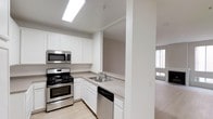 This single-floor space showcases a well-appointed kitchen designed for both functionality and visual appeal. The kitchen is outfitted with a full suite of stainless steel appliances, including a gas range with oven, an over-the-range microwave, and a dishwasher. Crisp white cabinetry lines the walls, providing generous storage above and below the counter level. The light-toned countertops complement the clean, neutral color palette throughout. A double-basin sink is positioned beneath upper cabinets, and a skylight overhead floods the kitchen with bright, even illumination, reducing the need for artificial lighting during the day. Light wood-style flooring runs through the kitchen, adding warmth to the otherwise modern aesthetic. A structural column subtly defines the boundary between the kitchen and the adjacent living area, which is visible in the background. That adjoining space features neutral carpeting and a window that allows natural light to filter in, contributing to an open and airy atmosphere. The overall layout promotes a connected, flowing feel between the kitchen and living spaces.