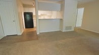 This single-floor residential unit offers a practical and functional layout designed for comfortable everyday living. The living area is finished with wall-to-wall neutral-toned carpeting, creating a warm and understated foundation for the space. A smooth transition separates the carpeted living zone from the kitchen, which features hardwood-style flooring for easy maintenance.

The kitchen is well-appointed with crisp white cabinetry providing ample storage, a stainless-style sink, and a peninsula-style breakfast bar with a countertop surface that doubles as a casual dining or prep area. The bar opening connects the kitchen to the living space, enhancing the open-concept feel and encouraging interaction between the two zones. A full-size black refrigerator anchors one end of the kitchen, complementing the clean white cabinetry throughout.

Recessed and track lighting illuminate the kitchen area, ensuring bright and functional task lighting. A floor-level HVAC vent is integrated into the layout, contributing to year-round climate comfort. The neutral wall tones and understated finishes throughout the unit create a cohesive, move-in-ready atmosphere that provides a versatile backdrop for a variety of interior styles.