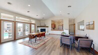 Located at 3314 Kennsington Ct in Manhattan, Kansas, this single-story community amenity space for Cambridge Apartments offers a thoughtfully designed environment for residents to gather, work, and stay active. The entry foyer greets visitors with soaring ceilings, a statement geometric pendant light, and a set of wood-framed glass doors that flood the space with natural light. Warm-toned luxury vinyl plank flooring flows throughout, creating a cohesive and inviting atmosphere. The reception and lounge area features tufted leather accent chairs, a dedicated workspace, and curated wall art, establishing a professional yet comfortable welcome zone. The expansive open gathering room serves as the heart of the space, anchored by a classic brick fireplace with a wood mantel and surrounded by large French doors and transom windows that offer views of the outdoor patio area. A dining table and area rug define this social zone, which connects seamlessly to a pass-through kitchen featuring granite countertops, warm wood cabinetry, and a chandelier overhead. Rounding out the amenities is a well-equipped fitness center with cardio machines and strength training equipment, making this a comprehensive and functional community hub.