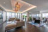 This elegantly designed single-floor amenity space offers a seamless blend of modern sophistication and inviting comfort. The open-plan layout flows effortlessly between distinct functional zones, all unified by rich dark hardwood flooring, recessed lighting, and a refined neutral palette accented with bold pops of color.

The main lounge area features generous sectional seating, sheer curtain dividers, and lush indoor plants that lend an airy, organic quality to the space. Floor-to-ceiling windows and sliding glass doors bathe the interior in abundant natural light while framing impressive panoramic city views and providing access to an outdoor terrace.

A dedicated media lounge offers plush sectional sofas arranged around a large built-in entertainment wall with a flat-screen display, framed by architectural pendant lighting with an industrial-modern character. Cove lighting along a tray ceiling adds warmth and dimension.

The kitchen and bar area is a standout feature, showcasing a long white quartz countertop lined with sleek black-and-gold bar stools, warm wood cabinetry, a textured stone mosaic backsplash, stainless steel appliances, and a striking multi-arm brass chandelier overhead.

Intimate dining areas with round stone-topped tables and upholstered chairs are positioned near large windows, offering elevated city views with sheer, ombre drapery providing a soft, sophisticated finishing touch throughout.