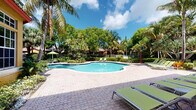 This inviting outdoor amenity area embodies a classic Florida resort aesthetic, centered around a generously sized freeform swimming pool with crystal-clear blue water. The pool deck is finished in decorative paver stonework in a distinctive hexagonal pattern, providing ample space for sunbathing and relaxation. A collection of green cushioned chaise lounges and umbrella-shaded seating is thoughtfully arranged around the pool, offering both sunny and shaded options for guests.

Surrounding the pool, lush tropical landscaping creates a vibrant, secluded atmosphere, with towering palm trees, mature shade trees, and well-maintained tropical shrubs forming a natural green canopy. The verdant backdrop enhances the sense of privacy and escape.

Adjacent to the pool is a Mediterranean-style clubhouse building featuring warm yellow stucco exterior walls, a red tile roof, and large multi-pane windows that allow natural light to fill the interior. Decorative stone columns frame a covered outdoor terrace, which is furnished with a spacious sectional sofa in bold orange upholstery, creating a comfortable and stylish al fresco lounge. The paved entryway leads directly to the building's main entrance. Together, these elements create a cohesive, resort-inspired setting ideal for leisure and outdoor gathering.