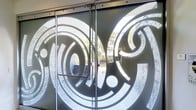 This single-floor space makes an immediate architectural statement through its entrance, where a pair of large frosted glass sliding doors serve as the visual centerpiece. The doors are emblazoned with bold, intricate circular and spiral graphic motifs — a striking design choice that blends artistry with functionality. The frosted treatment of the glass allows natural light to filter through while maintaining privacy, and the contrast between the opaque patterned sections and the clear glass creates a dynamic visual effect.

The entrance doors are framed by a sleek stainless steel hardware system, including a top-mounted sliding track and push-pull handles, reflecting a contemporary and polished aesthetic. Through the transparent portions of the glass, glimpses of a landscaped exterior are visible, suggesting that natural surroundings and greenery are a complementary feature of the overall environment. Stone or textured wall cladding is also partially visible beyond the entrance, hinting at a refined material palette used throughout the property.

The overall ambiance at the entry point communicates a professional, design-forward sensibility — ideal for a commercial, cultural, or hospitality setting. The combination of decorative glass artistry, quality hardware finishes, and the integration of natural exterior views establishes a welcoming yet sophisticated tone for the space within.