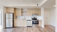 This single-floor unit showcases a cohesive modern design with light wood-tone flooring that runs consistently throughout the space. The kitchen is a standout feature, outfitted with flat-front cabinetry in a warm natural wood finish, open shelving, and a white subway tile backsplash that adds texture to the clean aesthetic. Stainless steel appliances include a top-mount refrigerator, electric range with hood, and dishwasher, all paired with light-toned countertops and an undermount sink. Natural light filters into the kitchen area through a nearby window, brightening the space considerably.

The bedroom is bright and airy, benefiting from a large picture window that frames neighborhood views and allows generous daylight. The white walls and light flooring contribute to an open, uncluttered feel.

The bathroom maintains the unit's modern sensibility with a white soaking tub and shower combination, a navy blue vanity with an undermount sink, a large mirror, and in-unit stacked washer and dryer conveniently integrated into the space. Recessed lighting and light flooring keep the bathroom feeling clean and well-lit. Overall, the unit presents a fresh, contemporary interior with consistent finishes throughout.