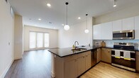 This single-floor residence showcases a thoughtfully designed open-concept layout that seamlessly connects the kitchen and living areas. The kitchen is a standout feature, equipped with stainless steel appliances including a French door refrigerator, electric range, and built-in microwave. Two-tone cabinetry pairs warm wood lower cabinets with crisp white upper cabinets, complemented by dark granite countertops and a textured tile backsplash. Globe pendant lights hang above the central island, which includes a sink and offers additional prep space. Recessed lighting and track lighting provide ample illumination throughout.

The living area extends naturally from the kitchen, featuring wood-look flooring and French doors that welcome natural light. Bedrooms are finished with plush carpeting, neutral walls, and windows fitted with blinds, creating a calm and restful atmosphere.

The bathroom is well-equipped with an extended vanity featuring light wood cabinetry and a white countertop, a wide mirror with wall-mounted lighting, wood-look vinyl flooring, and a tiled tub-shower combination. Throughout the unit, a consistent palette of warm neutrals and modern materials creates a cohesive, contemporary aesthetic.