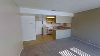 This single-story space features an open-concept design that connects the kitchen and living area seamlessly. The kitchen is equipped with white appliances, including a freestanding gas range, oven, and a full-size refrigerator. Cabinetry lines the upper and lower walls, providing ample storage, while an under-cabinet light fixture illuminates the workspace. A built-in breakfast bar with a laminate countertop extends into the living area, offering a convenient dining or casual workspace surface. The kitchen is finished with tile flooring, which transitions into neutral wall-to-wall carpeting in the spacious living area. A textured popcorn ceiling and a structural beam detail are consistent throughout both spaces. Recessed HVAC venting is visible overhead. The living area benefits from natural light entering through a window, casting sunlit patterns across the carpet. The overall color palette is neutral, with white walls, light cabinetry, and warm-toned flooring, creating a clean and functional environment suitable for everyday living.