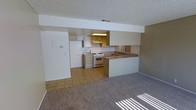 This single-story space features an open-concept design that connects the kitchen and living area seamlessly. The kitchen is equipped with white appliances, including a freestanding gas range, oven, and a full-size refrigerator. Cabinetry lines the upper and lower walls, providing ample storage, while an under-cabinet light fixture illuminates the workspace. A built-in breakfast bar with a laminate countertop extends into the living area, offering a convenient dining or casual workspace surface. The kitchen is finished with tile flooring, which transitions into neutral wall-to-wall carpeting in the spacious living area. A textured popcorn ceiling and a structural beam detail are consistent throughout both spaces. Recessed HVAC venting is visible overhead. The living area benefits from natural light entering through a window, casting sunlit patterns across the carpet. The overall color palette is neutral, with white walls, light cabinetry, and warm-toned flooring, creating a clean and functional environment suitable for everyday living.
