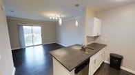 This single-floor space showcases an open-concept layout that integrates the kitchen and living areas into one cohesive environment. The kitchen is appointed with crisp white cabinetry, dark granite-style countertops, and a peninsula island that doubles as a functional workspace and casual dining surface. A stainless steel sink is embedded into the island, complemented by a sleek faucet and a built-in dishwasher beneath. Subway tile backsplash adds a clean, classic touch behind the upper cabinetry. Pendant lighting fixtures hang above the island, providing focused task lighting while enhancing the overall aesthetic alongside a coordinating track light fixture nearby. Crown molding runs along the ceiling, lending a refined architectural detail throughout the space. The living area extends naturally from the kitchen, featuring dark hardwood-style flooring that grounds the interior with warmth and depth. A sliding glass door at the far end of the living space floods the interior with natural light and provides access to an outdoor area, offering views of the surrounding landscape. The neutral wall tones and open layout create a bright, versatile atmosphere throughout.