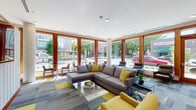 Located on NE Martin Luther King Jr Blvd in Portland's vibrant NE neighborhood, this single-floor amenity space offers a well-appointed and thoughtfully designed environment. The centerpiece is a spacious lounge area bathed in natural light from expansive floor-to-ceiling windows framed in warm wood trim, providing open views of the surrounding streetscape and mature trees. The lounge is furnished with generous sectional sofas and accent chairs in grey and yellow tones, complemented by a colorful modular carpet in grey and yellow patterns that adds energy to the space. A textured white feature wall with a mounted flat-screen display serves as a focal point, with linear accent lighting adding a modern touch to the interior. The kitchenette is outfitted with light wood cabinetry, a stainless steel refrigerator, microwave, sink, and open glass shelving with white subway tile backsplash — ideal for casual gatherings. The fitness center is well-equipped with treadmills, an elliptical, a stationary bike, and a weight bench, all set on durable rubber flooring with a large wall mirror enhancing the sense of space. Recessed lighting throughout ensures a bright, welcoming atmosphere across all areas.