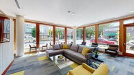 Located on NE Martin Luther King Jr Blvd in Portland's vibrant NE neighborhood, this single-floor amenity space offers a well-appointed and thoughtfully designed environment. The centerpiece is a spacious lounge area bathed in natural light from expansive floor-to-ceiling windows framed in warm wood trim, providing open views of the surrounding streetscape and mature trees. The lounge is furnished with generous sectional sofas and accent chairs in grey and yellow tones, complemented by a colorful modular carpet in grey and yellow patterns that adds energy to the space. A textured white feature wall with a mounted flat-screen display serves as a focal point, with linear accent lighting adding a modern touch to the interior. The kitchenette is outfitted with light wood cabinetry, a stainless steel refrigerator, microwave, sink, and open glass shelving with white subway tile backsplash — ideal for casual gatherings. The fitness center is well-equipped with treadmills, an elliptical, a stationary bike, and a weight bench, all set on durable rubber flooring with a large wall mirror enhancing the sense of space. Recessed lighting throughout ensures a bright, welcoming atmosphere across all areas.