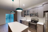 Located at 1550 Perimeter Pkwy NW in Huntsville, Alabama, this thoughtfully designed one-bedroom, one-bathroom residence makes excellent use of its compact footprint. The open-concept kitchen and living area serves as the heart of the home, featuring a striking blue kitchen island topped with white quartz countertops and illuminated by bold teal pendant lights. Sleek gray wood-grain cabinetry — including glass-front upper cabinets — pairs with stainless steel appliances, including a range, built-in microwave, dishwasher, and refrigerator, to create a fully equipped and visually cohesive cooking space. Light wood-look flooring runs throughout, lending warmth and continuity to the entire residence. The bedroom is a well-proportioned retreat with a large window that invites natural light and features elegant draping curtains. Two closets provide ample storage. The bathroom is finished with a wood-toned vanity, white quartz countertop, and an illuminated mirror, complemented by built-in open shelving. A stacked washer and dryer unit is conveniently integrated into the bathroom area, adding everyday practicality. Clean lines, neutral tones, and modern fixtures throughout create a polished, move-in-ready atmosphere.