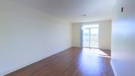 This single-floor space offers a clean, open layout defined by rich, dark hardwood flooring that contrasts beautifully with the crisp white walls and ceiling. The interior is bathed in natural light, primarily sourced from a large glass door at the far end of the main living area. This door opens onto a private balcony enclosed by a railing, offering views of the surrounding greenery and sky beyond — a notable feature that brings the outdoors in and enhances the sense of openness within the space.

The walls are finished in a neutral off-white tone, providing a versatile backdrop suitable for a variety of interior styles. Ceiling-mounted HVAC vents are discreetly integrated into the ceiling, ensuring climate control without disrupting the clean aesthetic. The absence of interior clutter allows the architectural simplicity to stand out, highlighting the well-maintained flooring and the generous proportions of the room.

Overall, the space conveys a light, airy ambiance with a modern yet understated character. The combination of natural light, hardwood flooring, and outdoor access creates a welcoming environment that feels both functional and comfortable.