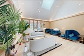 This single-floor living room lounge offers a refined and welcoming atmosphere, characterized by a striking vaulted ceiling with a central skylight that floods the space with natural light. Warm, light wood paneling lines the walls, complementing the pale hardwood floors to create a cohesive Scandinavian-inspired aesthetic throughout.

The room is thoughtfully arranged with distinct seating zones. A pair of softly curved light gray sofas anchor a central lounge area, accented by a low round coffee table and layered area rugs. Vibrant teal high-back sofas and rounded navy accent chairs provide rich pops of color against the neutral backdrop, adding depth and visual interest to the overall design.

Floor-to-ceiling wood-framed windows along one wall offer generous views of an outdoor terrace and courtyard, further enhancing the sense of openness. Sculptural mushroom-cap floor lamps are positioned throughout, contributing warm ambient lighting alongside recessed ceiling fixtures and subtle cove lighting.

A low credenza along the back wall displays curated books and decorative objects beneath a bold abstract painting in warm pinks and blues, serving as a focal point. Lush tropical and leafy plants are placed throughout, bringing vitality and a natural, organic touch to the contemporary interior. The result is a bright, elegant, and inviting space ideal for relaxed gathering and conversation.