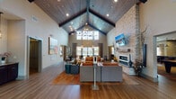 The Reserve At The Boulevard's clubhouse at 1 Reserve Drive in Yaphank, New York, is a well-appointed community amenity center designed to offer residents a full range of social and wellness facilities under one roof. The grand entrance sets the tone immediately, with soaring double-height ceilings clad in warm wood paneling, exposed dark beams, and a striking vaulted roofline visible through a large interior clerestory window. Floor-to-ceiling glass entry doors open into a spacious lounge area featuring hardwood flooring, plush seating arrangements, a dramatic stone fireplace with a mounted flat-screen television, and a sculptural chandelier. Natural light flows generously throughout the main gathering space. Adjacent to the lounge is a kitchenette outfitted with granite countertops, dark cabinetry, a mosaic tile backsplash, and pendant lighting, with views toward an outdoor pool area. A billiards and game room provides casual seating and billiard-themed artwork against textured walls. The fitness wing includes a cardio room lined with Matrix equipment and large windows, a dedicated spin studio with cycling bikes and colorful accent walls, and a bright yoga and movement studio with mirrored walls, light hardwood flooring, and a ceiling fan. A package locker station and closet storage round out the practical amenities of this thoughtfully designed facility.