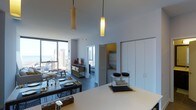 This single-floor high-rise unit offers an open-concept layout that seamlessly integrates the living, dining, and kitchen areas into one cohesive space. Soaring ceilings amplify the sense of volume throughout, while dramatic floor-to-ceiling windows flood the interior with abundant natural light and frame expansive views of the surrounding city skyline and the ocean beyond.

The kitchen features a clean, minimalist island with a bright white countertop that anchors the space and serves as a natural gathering point. Cylindrical pendant lights in warm tones hang at varying heights above, adding character and functional illumination. The dining area accommodates a dark wood table with seating, positioned to take advantage of the panoramic views.

Adjacent to the main living area, a large built-in wardrobe with floor-to-ceiling paneled doors provides generous storage while maintaining the space's streamlined aesthetic. A dedicated sleeping area is partially enclosed by a louvered door, offering a degree of privacy within the open-plan arrangement. Accent pieces in yellow add warmth to the otherwise neutral grey and white palette, contributing to a modern, polished ambiance throughout.