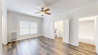 Located at 849 S Gramercy Dr in the heart of Los Angeles, this single-floor unit offers a thoughtfully arranged layout comprising a living room, a combined dining room and kitchen, two hallways, and a full bathroom. The living room is a bright, open space defined by warm hardwood-style flooring that runs throughout the unit. Charming decorative wall molding and arched panel detailing lend the space a classic architectural character, while a ceiling fan provides both comfort and visual interest. Natural light filters in through the windows, enhancing the clean, neutral color palette of the white walls. The combined dining and kitchen area features a galley-style layout equipped with white cabinetry, a gas range, a stainless steel refrigerator, and a sink with countertop space — efficiently designed for everyday use. A window at the end of the kitchen brings in additional natural light. The bathroom is accessible via the hallway and includes a full bathtub with a classic black tile border accent, a vanity with a wood-toned cabinet, a toilet, and a window that allows for natural ventilation. The overall ambiance of the unit is clean, practical, and inviting, with timeless design elements that add character throughout.