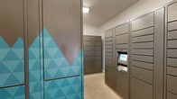 Located at 300 NC-146 in Asheville, NC, this single-floor mail and package room is purpose-built to provide a streamlined, secure solution for receiving and storing deliveries. The room is lined on multiple sides with rows of gray metal parcel lockers of varying sizes, accommodating everything from small envelopes to larger parcels. A centrally positioned smart locker kiosk features a digital touchscreen interface, allowing recipients to retrieve packages using unique access codes, ensuring both security and convenience.

Traditional gray metal mailboxes are also present, offering dedicated compartments for standard mail alongside the parcel lockers. The layout creates a narrow corridor between banks of lockers, making efficient use of the available space while maintaining clear accessibility throughout. A standout design element is a bold teal geometric-patterned panel near the entrance, adding a modern, visual accent that contrasts with the neutral gray tones of the lockers and walls.

The flooring consists of light beige tile throughout, contributing to a clean and practical aesthetic. White walls and a textured ceiling with recessed lighting keep the space bright and functional. A white paneled door provides access to an adjacent area. Overall, the room is well-organized and designed to support high-volume mail and package handling efficiently.
