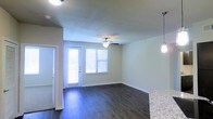 This single-floor residence offers a clean, functional layout centered around an open-concept living and dining area. The space features rich, dark hardwood-style flooring that runs throughout, providing a polished and cohesive aesthetic. Neutral-toned walls create a bright, welcoming atmosphere that pairs well with the natural light streaming in through multiple windows fitted with blinds. A ceiling fan with an integrated light fixture serves the main living area, ensuring both comfort and illumination. A glass-paneled exterior door offers additional light and direct access to the outside. Adjacent to the living area, a kitchen counter with a granite-style surface is visible, accented by a stylish pendant light hanging overhead, hinting at a well-appointed kitchen space nearby. The overall layout feels open and airy, with a seamless flow between the living, dining, and kitchen zones. The design balances practicality with modern touches, making the space approachable and comfortable. Clean lines, neutral finishes, and thoughtful lighting combine to create an inviting environment suited for everyday living.
