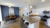 This 2-bedroom, 2-bathroom residence is designed with a modern open-concept layout that seamlessly connects the kitchen, dining, and living areas. The kitchen is a standout feature, boasting white cabinetry, dark countertops, and a large peninsula with bar seating — ideal for casual dining or entertaining. Pendant lighting above the peninsula adds a contemporary touch, while under-cabinet lighting enhances both functionality and ambiance. A dedicated sink and ample counter space make the kitchen as practical as it is attractive. The adjacent dining area features a bold navy accent wall that adds a pop of character and depth to the space, complementing the warm wood-look flooring that runs throughout. The open flow between rooms creates a sense of spaciousness and ease of movement. Natural light filters through the layout, contributing to a bright and welcoming atmosphere. The overall design aesthetic balances modern clean lines with warm, inviting details, making this residence a comfortable and visually appealing home. With two full bathrooms and two bedrooms, the floor plan is well-suited for roommates, guests, or those who desire extra space.