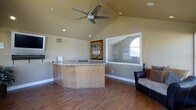 This single-floor space offers a welcoming and well-appointed interior defined by its vaulted ceilings that lend an airy, expansive feel to the main area. A central curved counter with a granite-style surface serves as a functional focal point, ideal as a reception desk or workstation, and is finished with warm wood paneling along its base. The flooring throughout is composed of rich hardwood, adding warmth and continuity to the space.

Recessed lighting is distributed across the ceiling, complemented by a contemporary ceiling fan that provides both comfort and style. A large interior window connects to an adjacent windowed alcove, allowing borrowed natural light to flow into the space and providing a sense of visual depth. An arched exterior window in the adjoining area further enhances the brightness of the overall environment.

The walls are finished in a neutral, warm beige tone that pairs harmoniously with the wood elements throughout. Seating is provided by a modern dark leather chair and an upholstered sofa, creating a comfortable area for guests or clients. Wall art and decorative dried botanicals add tasteful finishing touches. Ventilation is well integrated with visible floor and wall vents throughout, ensuring practical comfort in everyday use.
