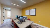 This single-floor business center is thoughtfully organized into two distinct functional zones, offering a well-rounded workspace environment. The computer lab occupies one section of the space and is equipped with multiple workstations, including iMacs and PC monitors, arranged along a long, neutral-toned communal desk. Red upholstered task chairs provide seating, while linear LED ceiling fixtures deliver consistent, even illumination throughout. A bold yellow accent wall serves as a visual focal point, complemented by two framed display panels. Carpeted tile flooring in a muted tone grounds the room with a professional aesthetic.

Separated by a frosted glass partition wall with a full-lite glass door, the adjacent meeting area offers a more intimate setting. A natural wood dining-style table is surrounded by mid-century modern chairs with upholstered seats. Striking black pendant lights hang overhead from exposed black structural beams, adding an industrial design element. A warm yellow accent wall continues the cohesive color theme, and a framed artwork piece and decorative console table enhance the space. A large window allows natural light to filter in, offering views of the surrounding exterior.