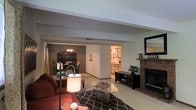 This single-floor residence offers a welcoming and well-connected layout designed for comfortable everyday living. The living room serves as the heart of the home, anchored by a classic brick fireplace that adds warmth and character to the space. A glass-top coffee table, plush seating, and a patterned area rug contribute to the room's inviting ambiance, while a floor lamp provides soft, layered lighting.

Structural beams along the ceiling add a subtle architectural detail that defines the living area while maintaining an open feel. A partial wall divides the space from an adjoining dining room, which features a chandelier and a dark wood hutch, lending the area a traditional and refined character.

A central hallway connects the main living areas to the rest of the home, offering a clear sightline through the space. Beyond it, a bright kitchen is visible, finished in light tones with ample counter space and cabinetry.

A media console with shelving sits opposite the fireplace, providing practical storage and display options. A decorative mirror on the fireplace wall adds depth to the room. Overall, the space presents a cohesive, homey aesthetic with warm neutral tones, traditional furnishings, and thoughtful details throughout.