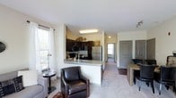 This single-floor unit showcases an open-concept design that seamlessly connects the living, dining, and kitchen areas, creating a cohesive and functional living environment. The kitchen is outfitted with dark espresso cabinetry, stainless steel appliances including a refrigerator and microwave, and a granite-topped peninsula that doubles as a breakfast bar. The peninsula provides additional prep space while acting as a natural divider between the kitchen and the living area. The living space features neutral carpeting and is illuminated by natural light streaming through a large window dressed with white blinds and curtains. A hallway visible from the main living area leads to additional rooms, each with windows that continue the theme of natural light throughout the unit. Gray doors add a contemporary accent to the otherwise warm, neutral color palette of beige and cream walls. The dining area sits adjacent to the living space, furnished with a wooden table and chairs. Overall, the layout offers a practical and inviting atmosphere, ideal for everyday living.