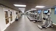 This fitness center is designed to accommodate a comprehensive workout experience within a single-floor layout. The space features a long, open floor plan lined with a generous row of cardio equipment, including treadmills, elliptical machines, and steppers, all arranged along one side for easy access and flow. Rubber flooring throughout provides both durability and comfort underfoot, making the space practical for a variety of exercise activities.

The ceiling is finished with a drop-tile grid and recessed fluorescent panel lighting, ensuring the room is evenly and brightly illuminated throughout. Windows along the far wall allow for natural light to supplement the artificial lighting, contributing to an open and energizing atmosphere. Large mirrors are positioned along the walls, visually expanding the space and allowing users to monitor their form during workouts.

Additional equipment, including stability balls and wall-mounted accessories, is stored along the perimeter, keeping the central floor area clear and accessible. The walls are painted in a soft, neutral tone, creating a clean and focused environment. Overall, the fitness center is thoughtfully arranged to support an efficient and comfortable exercise experience.