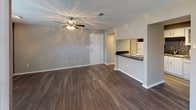 This single-story unit presents a thoughtfully arranged floor plan with a blend of practical features and comfortable finishes. The open living area is finished with rich wood-style luxury vinyl flooring, neutral gray walls, and a textured ceiling fitted with a ceiling fan and light fixture. Natural light enters through blinds-covered windows, and a sliding glass door with vertical blinds provides additional brightness. A standout feature of the living space is the full wall of built-in white shelving, offering generous storage and display options.

The layout flows into a secondary room, likely used as a bedroom or den, which features plush wall-to-wall carpet, a ceiling fan, and a window with natural light. The galley-style kitchen is efficiently designed with white cabinetry, dark laminate countertops, a black tile mosaic backsplash, white appliances including a range and refrigerator, and a stainless steel sink. A pass-through counter connects the kitchen to the main living area. The kitchen also leads to a utility area with additional storage.

The bathroom is clean and well-appointed, featuring a white vanity with dark countertop, a framed mirror with strip lighting, wood-style flooring, and a full tub-shower combination with white tile surround.