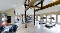This fitness center in Arlington offers a compelling blend of rustic character and modern functionality. The space is defined by striking exposed wooden beams and structural posts that rise toward vaulted ceilings, lending a warm, industrial-rustic aesthetic that sets it apart from a typical gym environment. The polished concrete floors are durable and easy to maintain, contributing to the open, airy feel of the room. Large windows on multiple walls flood the space with natural light, creating an inviting and energizing atmosphere for workouts. Full-length mirrored walls visually expand the room and provide practical utility for form-checking during exercise. The fitness center is well-appointed with a comprehensive selection of equipment, including treadmills, elliptical machines, and a multi-station strength training unit, ensuring a full range of cardio and resistance training options. A set of colorful medicine balls adds a functional training element to the floor space. Recessed lighting supplements the natural light, ensuring the space remains bright and well-illuminated at all times. The overall ambiance is both motivating and comfortable, making this fitness center a standout amenity.