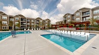 This outdoor amenity space is the centerpiece of a multi-story residential community, offering a thoughtfully designed retreat for relaxation and recreation. The complex features two distinct swimming pools: a large rectangular lap-style pool and a separate shallow wading pool, both finished with clean blue tile and crystal-clear water. Surrounding the pools is a generously sized concrete sundeck lined with white lounge chairs, providing ample space for sunbathing and outdoor leisure. A decorative stone water feature wall serves as a striking architectural divider between the two pool areas, adding visual depth and character to the space. A classic wood pergola with red accent planters and seating anchors one corner of the deck, creating a shaded gathering spot. Additional amenities include outdoor dining areas with tables and umbrellas, grilling stations, and vibrant red accent furniture that adds a bold pop of color throughout. The entire pool area is enclosed by wrought-iron fencing, ensuring privacy and security. The surrounding multi-story residential buildings, clad in warm brown and gray tones, frame the space and contribute to a cohesive, community-oriented atmosphere. Mature trees and landscaping add a welcoming touch, while open skies overhead enhance the bright, airy ambiance of this expansive outdoor living area.