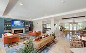 Located on Nuckols Road in Glen Allen, Virginia, this expansive single-floor community amenity center offers a thoughtfully designed and generously proportioned gathering space. Light hardwood flooring flows throughout, unifying the open-concept layout and complementing the bright, airy atmosphere created by abundant natural light from large windows overlooking outdoor courtyards and landscaped grounds.

The centerpiece lounge features a striking teal built-in wall unit with integrated shelving, a modern electric fireplace, and a mounted TV, flanked by vibrant orange sofas atop a colorful area rug. Adjacent to the lounge, a well-appointed coffee bar — branded as Triton Coffee House — showcases custom blue cabinetry, granite countertops, mosaic tile backsplash, and stainless steel appliances, with a large island providing additional seating.

The open dining area accommodates multiple seating arrangements beneath pendant lighting, with white columns and greenery walls adding architectural interest. A game room with a billiards table and a cozy private meeting nook — featuring a teal geometric accent wall, banquette seating, and garden views — offer versatile spaces for socializing or focused gatherings. Completing the amenity suite is a dedicated private theater room with tiered leather recliner seating, moody dark walls, navy drapes, and classic movie poster décor.