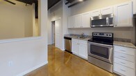 This single-floor loft-style unit combines industrial character with modern finishes throughout. The open living area makes a striking first impression with its richly stained, polished concrete floors that reflect light across the space, while the dark exposed beams and black-painted ceiling with recessed track lighting overhead add a distinct industrial aesthetic. A row of windows along one wall floods the main living area with natural light, offering views of surrounding greenery and creating an airy, open atmosphere.

The kitchen is well-appointed with crisp white shaker cabinetry, granite countertops, and a full suite of stainless steel appliances including a refrigerator, electric range, built-in microwave, and dishwasher. The layout provides generous counter and storage space, and flows seamlessly into the main living area, making it ideal for both everyday living and entertaining.

The bathroom continues the unit's consistent design language, featuring polished concrete floors, a white vanity with granite countertop, an undermount sink, and a tub/shower combination. Clean white walls and chrome fixtures give the space a tidy, functional feel.

Practicality is further enhanced by a dedicated in-unit laundry closet housing a stacked washer and dryer. Thoughtful HVAC venting throughout ensures year-round comfort in this uniquely styled space.