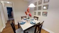 Located at 321 Harbor Drive in Claymont, Delaware, this single-floor home offers a thoughtfully designed layout with a warm and inviting ambiance throughout. The dining area is a standout feature, showcasing rich hardwood flooring that adds warmth and character to the space. A classic pendant light hangs above a light-toned rectangular dining table surrounded by dark wood cross-back chairs, creating an elegant yet comfortable setting for gathering and entertaining. The walls are adorned with an artfully arranged grid of square framed mirrors, adding depth and visual interest to the room while reflecting light to brighten the space. Neutral-toned walls provide a clean, versatile backdrop that complements the décor seamlessly. A hallway visible from the dining area hints at additional living spaces beyond, suggesting a functional and connected floor plan. The overall aesthetic is polished and welcoming, blending classic design elements with contemporary touches to create a home that feels both stylish and livable.