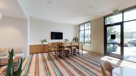 This single-floor space offers a clean, contemporary design characterized by crisp white walls, recessed lighting, and a smooth white ceiling that enhances the sense of openness. The centerpiece of the room is a substantial bar-height dining or meeting table surrounded by cushioned high-back chairs, making it well-suited for both casual gatherings and collaborative sessions. A wall-mounted flat-screen television is positioned above a warm wood-toned credenza, which provides additional storage and display space, accented by small potted greenery.

A striking floor-to-ceiling window along one wall floods the interior with abundant natural light and frames views of palm trees and the surrounding outdoor environment, creating a seamless connection between indoors and out. The dark-framed window adds a bold architectural contrast to the neutral walls.

Anchoring the space is a vibrant multicolored striped area rug in tones of red, teal, blue, and cream, introducing a lively visual element that complements the otherwise understated palette. A decorative wheel accent piece adds a subtle industrial touch. Overall, the space balances modern minimalism with warm, inviting details.