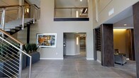 Located at Bishops, this impressive lobby space immediately commands attention with its soaring double-height ceilings and open, airy design. Large-format floor tiles in a warm grey tone anchor the space and contribute to a polished, professional aesthetic. The dramatic vertical volume is accentuated by a mezzanine-level walkway above, enclosed by sleek cable railings with a dark wood top rail, offering a sense of architectural depth and connectivity between levels. A carefully curated piece of framed abstract artwork adorns one of the neutral-toned walls, adding a creative focal point and warmth to the otherwise contemporary palette. Lush potted plants introduce a natural element, softening the crisp lines of the modern interior. Recessed and linear lighting fixtures illuminate the space effectively, highlighting the clean architectural lines throughout. A corridor leads further into the building, lined with frosted glass panels that allow light to filter through while maintaining privacy. On the opposite side, a textured dark accent wall adds contrast and visual interest. The overall ambiance is refined and welcoming, making a strong first impression suited to a professional or mixed-use environment.