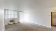 This single-floor unit is thoughtfully laid out to maximize functionality across its compact footprint. The living area serves as the central gathering space, featuring neutral wall-to-wall carpeting, clean white walls, and a large window dressed with vertical blinds that allow generous natural light to filter in. A wall-mounted air conditioning unit and baseboard heating ensure year-round comfort. The open feel of the living space is enhanced by its clean sightlines and uncluttered design.

The galley-style kitchen runs along a single corridor and is equipped with medium-toned wood cabinetry featuring brushed metal hardware, light countertops with an integrated sink, and white appliances including a refrigerator and electric range. Light tile flooring grounds the space practically.

The bedroom offers a comfortable footprint with carpeted flooring, a window with vertical blinds, and access to a dedicated closet with built-in shelving. The full bathroom includes a wood-toned vanity with light countertops, tile accents, a toilet, and a tub-shower combination. Wood-grain interior doors carry a consistent design element throughout the unit.