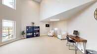 Located at 6050 Hendry Dr in North Chesterfield, VA, this single-floor community space offers a versatile and welcoming environment designed for both relaxation and productivity. The room immediately impresses with its dramatically soaring ceilings, which are accentuated by clerestory windows positioned high on the upper walls, flooding the interior with abundant natural light. Large, ground-level windows along one wall provide sweeping views of the surrounding exterior, further enhancing the bright and airy ambiance.

The space is thoughtfully arranged into distinct functional zones. A cozy reading and lounge area features two patterned accent chairs positioned around a side table, complemented by a well-stocked navy blue bookshelf that adds both color and character. A mounted display panel is conveniently accessible nearby. The business center area includes two wooden hairpin-leg desks, each equipped with a computer monitor and a sleek black chair, making it a practical resource for residents. A small round café-style table with two navy chairs occupies a quieter corner, ideal for casual meetings or conversation.

Neutral, warm-toned walls and plush wall-to-wall carpeting in a soft beige create a cohesive and calming atmosphere throughout. Potted plants add touches of greenery, contributing to the space's welcoming character. The open-plan layout and generous vertical volume make this a distinctive and functional communal amenity.