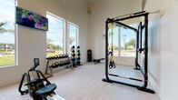 This two-story fitness facility is designed with a clean, modern aesthetic, characterized by crisp white walls, light hardwood-style flooring, and soaring double-height ceilings on the main level. Tall columns punctuate the open floor plan, adding architectural structure while maintaining an expansive, airy atmosphere. Linear LED lighting complements the abundant natural light that streams through large windows, many of which frame views of palm trees and the surrounding outdoor landscape.

The main level is comprehensively equipped for a wide range of training disciplines. Cardio equipment, including treadmills, ellipticals, and rowing machines, is arranged near the windows, allowing users to exercise with outdoor views and wall-mounted televisions for entertainment. Strength training is well-represented with cable machines, a Smith machine, weight bench, leg extension machine, and a full dumbbell and kettlebell rack. A functional training area features a TRX suspension rig, medicine balls, and additional free weights. Built-in shelving and a water station add practical convenience.

A staircase leads to the upper level, which opens to the main floor below via a cable-rail mezzanine balcony. This secondary level houses a dedicated cycling studio fitted with stationary bikes and a wall-mounted television, offering a more intimate workout environment. Together, both floors deliver a well-rounded, professionally appointed fitness experience.