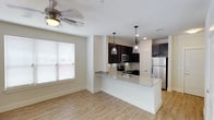This single-floor residence offers a thoughtfully designed open-concept layout that seamlessly connects the living and kitchen areas, creating a bright and functional living environment. The kitchen is a standout feature, showcasing dark espresso cabinetry that provides a striking contrast against the light granite countertops. A peninsula with an integrated sink and additional counter space serves as both a prep area and a casual gathering spot. Stainless steel appliances, including a refrigerator, microwave, and range, complete the kitchen's modern aesthetic. Overhead, two elegant pendant lights hang above the peninsula, adding a touch of sophistication and focused task lighting. The living area benefits from natural light streaming through a large window fitted with horizontal blinds, and a ceiling fan ensures year-round comfort. Light-toned hardwood-style flooring runs throughout the space, lending warmth and visual continuity. Crisp white walls and clean baseboards contribute to the overall fresh, contemporary feel of the interior. A paneled door visible in the background hints at additional private spaces within the residence. The overall ambiance is clean, modern, and move-in ready.