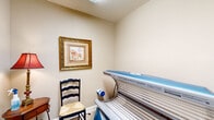 This single-room tanning space at 3300 Stadium Dr in Phenix City, Alabama, offers a compact and functional environment designed specifically for tanning services. The room is finished with warm, neutral beige walls that create a calm and welcoming atmosphere, complemented by light tile flooring laid in a diagonal pattern for a clean, polished look.

The centerpiece of the room is a Sunvision 24S tanning bed, a commercial-grade unit equipped with multiple tanning lamps beneath a blue-accented canopy. A wall-mounted paper towel dispenser and sanitizing supplies are conveniently accessible, reflecting an attention to cleanliness and client care.

Decorative touches elevate the room's ambiance beyond purely utilitarian — a gold-framed equestrian artwork adorns the wall, and a classic ladder-back chair with a woven rush seat provides a waiting area. A decorative lamp with a rich red shade and a lean-against-wall mirror add warmth and practicality to the space.

The room connects to a hallway through a white-trimmed doorway, hinting at its placement within a larger facility. Natural light is introduced subtly through a skylight above, brightening the interior without disrupting the tanning environment. Overall, the space balances professional functionality with tasteful décor.