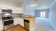 This single-floor, 2-bedroom, 2-bathroom home offers a well-designed open-concept layout that seamlessly connects the kitchen and living areas. The kitchen features white cabinetry, granite-look countertops, stainless steel appliances including a range and dishwasher, and a double-basin sink. A peninsula counter extends into the space, creating a natural divide between the kitchen and the adjoining living area while maintaining an open, airy feel. Track lighting above the kitchen provides focused illumination, complementing the overall brightness of the space. The living area is finished with plush carpeting and benefits from natural light streaming through a set of glass doors, which likely lead to an outdoor area. Crown molding runs along the ceiling throughout, adding a refined architectural touch. The soft, neutral color palette of light blue-gray walls and white trim creates a clean, cohesive aesthetic across the space. The wood-look flooring in the kitchen transitions smoothly into the carpeted living area, offering both practicality and comfort throughout the home.