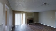 Located in Tyler, Texas, this single-story space offers a comfortable and functional layout with notable design details throughout. The main living area serves as the heart of the space, showcasing rich, dark hardwood-style flooring that contrasts elegantly with the warm, neutral-toned walls. A classic fireplace with a stone surround and a white mantel serves as a natural focal point, complemented by a framed accent wall above that adds a touch of architectural character. A large sliding glass door dressed with vertical blinds allows an abundance of natural light to flood the room, creating a bright and welcoming atmosphere. The textured ceiling adds a traditional touch consistent with the overall aesthetic of the space. The open feel of the living area provides ample room for a variety of furniture arrangements, making it both versatile and inviting. Overall, the space combines practical design with warm, understated details, making it a well-suited environment for comfortable everyday living in the Tyler, TX area.