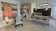 This single-floor residence offers a thoughtfully designed open-concept layout that seamlessly connects the living, dining, and kitchen areas. The living space features plush carpeting in warm neutral tones, complemented by decorative wall art and elegant wall sconces that add character and warmth to the room. A stylish accent chair with an ornate pattern anchors the seating area, creating an inviting atmosphere.

The kitchen is a standout feature, showcasing rich dark cabinetry that contrasts beautifully with light granite countertops. A peninsula with a built-in sink and sleek bar-height seating serves as both a functional workspace and a casual dining spot. A pendant light fixture hangs above, providing focused illumination and a contemporary design touch. Black appliances complete the kitchen's refined, cohesive aesthetic.

The bedroom, visible through a connecting hallway, continues the neutral color palette with soft bedding and subtle décor, offering a serene retreat. Recessed lighting throughout the space ensures consistent, well-distributed illumination across all areas. The overall ambiance is modern yet comfortable, with a cohesive design language that balances practicality and style throughout the entire floor plan.