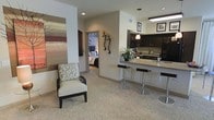 This single-floor residence offers a thoughtfully designed open-concept layout that seamlessly connects the living, dining, and kitchen areas. The living space features plush carpeting in warm neutral tones, complemented by decorative wall art and elegant wall sconces that add character and warmth to the room. A stylish accent chair with an ornate pattern anchors the seating area, creating an inviting atmosphere.

The kitchen is a standout feature, showcasing rich dark cabinetry that contrasts beautifully with light granite countertops. A peninsula with a built-in sink and sleek bar-height seating serves as both a functional workspace and a casual dining spot. A pendant light fixture hangs above, providing focused illumination and a contemporary design touch. Black appliances complete the kitchen's refined, cohesive aesthetic.

The bedroom, visible through a connecting hallway, continues the neutral color palette with soft bedding and subtle décor, offering a serene retreat. Recessed lighting throughout the space ensures consistent, well-distributed illumination across all areas. The overall ambiance is modern yet comfortable, with a cohesive design language that balances practicality and style throughout the entire floor plan.