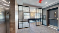 Located on the 5th floor at 975 Bonnel Ct in Union, NJ, this thoughtfully designed lounge space offers a refined and contemporary communal environment. The entry foyer sets an elegant tone with textured gray wall panels, plush carpet flooring, and a statement console table adorned with sculptural décor beneath a large round mirror — all positioned adjacent to elevator access.

Floor-to-ceiling steel-framed glass partitions divide the space into distinct functional zones while maintaining an open, transparent feel throughout. The living area features a generously sized sectional sofa with vibrant accent pillows, round upholstered ottomans, and decorative gold wall art set against soft gray walls with a tray ceiling detail. Natural light pours in through large windows, enhancing the airy ambiance.

A separate seating zone accommodates a marble-top round table with modern chairs and multiple wall-mounted flat-screen televisions, making it well-suited for both casual gatherings and media viewing. Colorful framed artwork adds personality throughout the space. Recessed lighting complements the natural light, ensuring a well-lit environment at all times. The overall design balances modern sophistication with a welcoming, comfortable atmosphere.