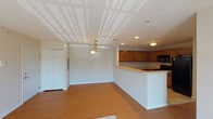 This single-floor unit offers a practical and functional layout designed to maximize living comfort. The main living area greets occupants upon entry, featuring warm hardwood-style flooring and a decorative chandelier overhead. A textured ceiling adds dimension to the space, while recessed lighting provides even illumination throughout. A half-wall peninsula visually separates the living area from the kitchen, creating an open-concept flow without sacrificing definition between spaces.

The kitchen is well-appointed with medium-toned wood cabinetry extending both above and below generous countertops, offering substantial storage and workspace. Black appliances, including a gas range, built-in microwave, and refrigerator, are integrated seamlessly. Tile flooring grounds the kitchen area practically.

The bedrooms are carpeted for comfort and feature neutral walls. One bedroom notably includes two side-by-side closets with bi-fold doors, while a second bedroom offers a single closet, each providing solid storage capacity. Natural light filters into the bedrooms through windows, with one room offering an exterior view of the surrounding landscape.

The bathroom includes a vanity with wood cabinetry, a single sink, a toilet, and a shower-tub combination with a curtain rod. Tile flooring completes the functional design.

The living area also features a sliding glass door that opens to an outdoor view, allowing natural light to brighten the carpeted lounge space.